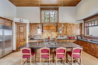 Fully equipped kitchen in The Lookout Home featuring custom wooden cabinetry, a large granite-topped island with four red barstools, and views of the pine trees outside.