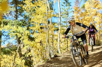 Two women mountain biking down a dirt path in a forest vibrant with autumn foliage.