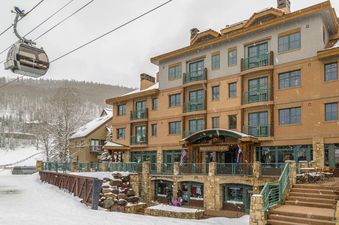 A multi-story building, the Inn at Lost Creek Junior Suite, is shown covered in snow during winter, with a gondola passing by and snowy mountains in the background.