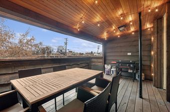 A covered outdoor dining area at The Loft features a long wooden table, wicker chairs, string lights, and a BBQ grill, offering a comfortable space for outdoor meals.