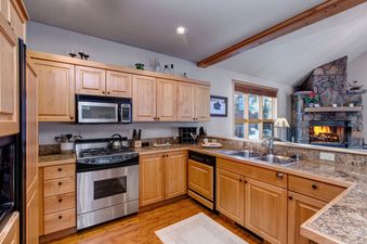A modern kitchen with wood cabinets, stainless steel appliances, and granite countertops. A fireplace is visible in the background.