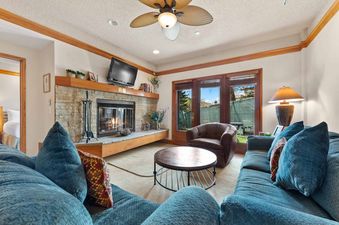 A cozy living room at The Charter C0405 features a stone fireplace, plush blue sofas, a round coffee table, and large windows with natural wood frames, offering a view of the outdoors.