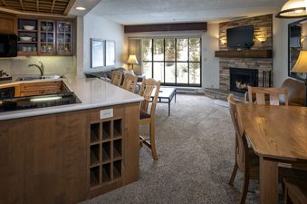 The interior of Building 4 One Bedroom Beaver Run Resort features a kitchen, dining area, and living room with a stone fireplace and a snowy outdoor view.