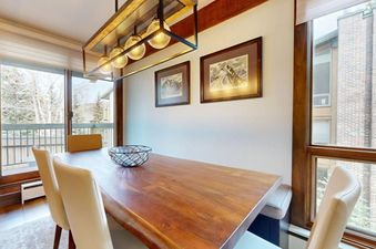 The dining area at Slope 101 features a wooden table with cream chairs, a unique chandelier, framed art, and large windows with a winter view.