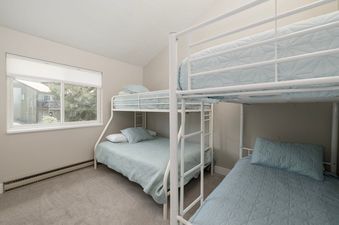 A cozy bedroom with two white metal bunk beds, each featuring light blue bedding and pillows, and a large window offering a view of other buildings and trees.