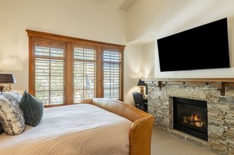 The bedroom in Deer Valley Lookout 26 features a comfortable bed with a leather headboard, a large flat-screen TV mounted above a stone fireplace, and windows with wooden shutters.