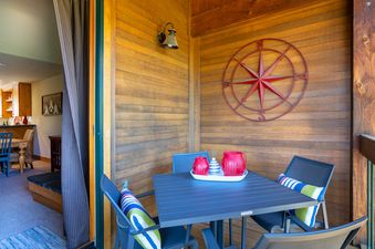 The private balcony features a dark gray dining table with matching chairs and colorful striped pillows, complemented by a wooden wall adorned with a red compass rose and a wall-mounted light fixture.