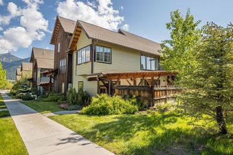 A charming townhouse with wooden siding, gabled roofs, and a front yard with green grass and shrubs, set against mountains.