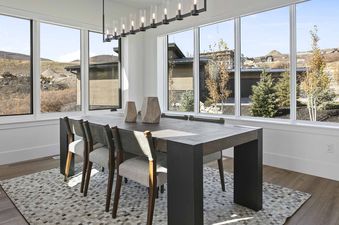 A modern dining room with a dark wooden table and light grey chairs, featuring large windows with views of hills and distant houses, and a patterned rug on a light wood floor.