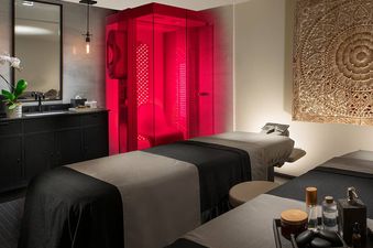 Two massage beds are positioned in a dimly lit spa room, with a red light therapy booth glowing intensely in the corner, and a carved wooden artwork on the far wall.