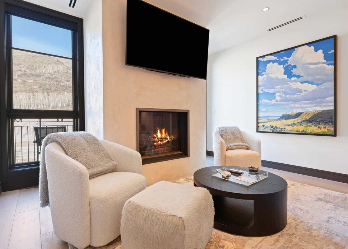 A cozy sitting area at Solaris PHCE with two cream armchairs, an ottoman, a dark wood coffee table, a fireplace with a TV above, and a large window with a mountain view.