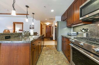 A modern kitchen with stainless steel appliances, dark wood cabinets, and granite countertops. The kitchen is open to a hallway with a door leading to a bedroom.