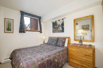 The bedroom in Meadow Ridge 28 Unit 6 features a queen-sized bed with a patterned comforter, nightstands, and a dresser with a mirror, all set against a neutral wall color with a window providing natural light.
