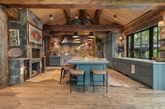 A spacious rustic kitchen with exposed wooden beams, a large stone fireplace, and blue cabinetry.