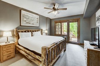 A rustic master bedroom in the Bear Paw Lodge 2 Bedroom with a log-frame king bed, wooden furniture, and outdoor access through glass doors leading to a green natural setting.