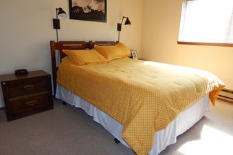 The image shows a cozy bedroom with a bed covered in a yellow patterned comforter, a wooden nightstand, and wall-mounted lamps.