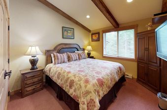 The primary bedroom at Terracehouse 64 features a large bed with a floral comforter, flanked by nightstands and lamps, with a window providing natural light and a wooden armoire with a television.