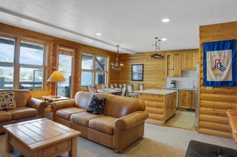 A cozy, rustic living and dining area with leather sofas, a wooden coffee table, a dining table set, and a kitchenette, all with views of a snowy exterior.