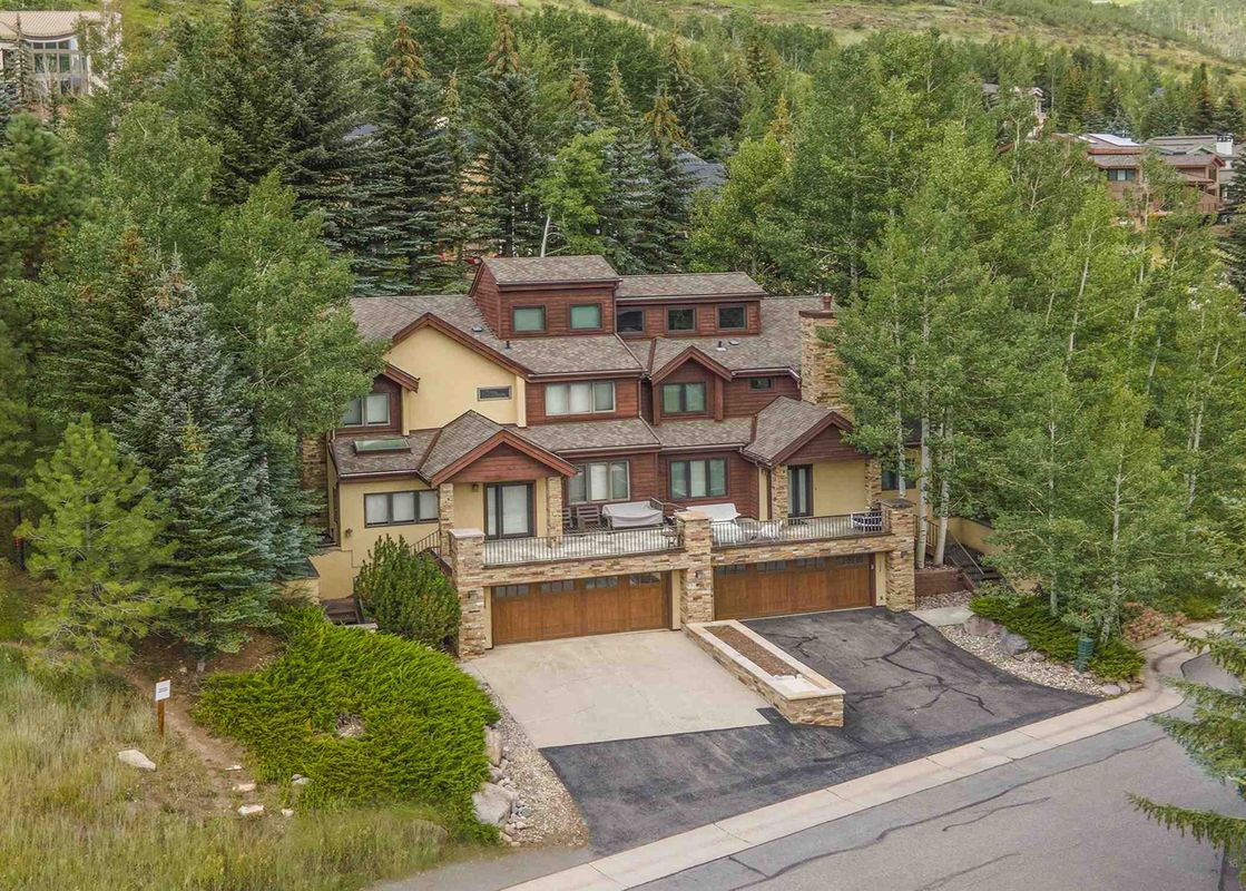 1175 A Casolar Del Norte, a three-story home, with three garage doors, a stone facade, a balcony, and a paved driveway, sits in the middle of a lush green forest.