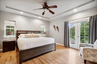 A serene bedroom at Suncrest Hideaway features a dark wood bed, light wood floors, a ceiling fan, and French doors opening to a forest view.