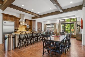 A wide view of the kitchen and dining area at Slopeside 111, featuring wood cabinets, stainless steel appliances, an island with bar stools, and a large dining table with chairs, all set against a backdrop of exposed wooden beams and large windows with gr