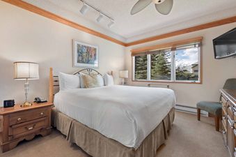 A spacious bedroom in The Charter C0405 with a king-size bed, white bedding, a wooden nightstand, a decorative lamp, and a large window with wooden blinds offering a view of evergreen trees and other buildings.