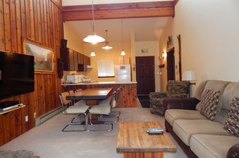 An inviting open-concept living area with wood-paneled walls, comfortable seating, a dining table, and a well-equipped kitchen.