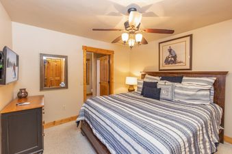 A cozy bedroom featuring a comfortable bed with striped bedding, a large wooden headboard, and a ceiling fan, with a dresser and mirror to the left and a doorway leading to a bathroom on the right.