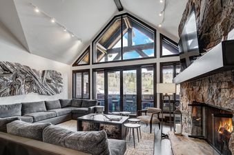 The spacious living room features a large grey sectional sofa, a dark coffee table, and an abstract metallic art piece. Floor-to-ceiling windows offer mountain views, while a stone fireplace adds warmth to the elegant space.