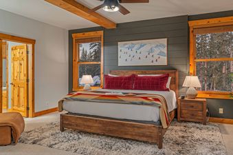 The bedroom at Starlit Pines Lodge features a king-size bed with a red velvet headboard and a plush blanket. The room has large windows with views of the snow-covered forest, and a wooden nightstand with a lamp. The room also features a cozy,  plush chair