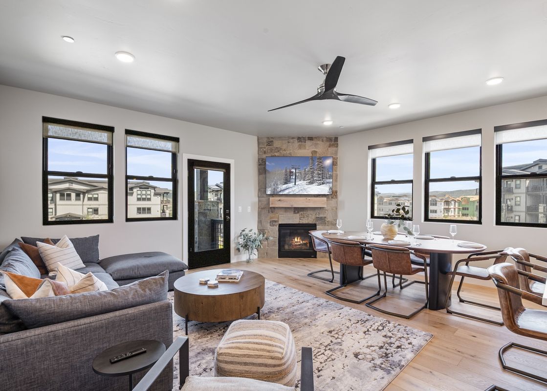A modern open-concept living and dining area with a sectional sofa, a fireplace with a TV above it, and a dining table with chairs, all illuminated by natural light from large windows.