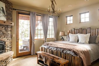 A rustic bedroom at Docs Holiday features a large bed with a wooden frame and headboard, a stone fireplace, and large windows with views of a hot tub and trees.