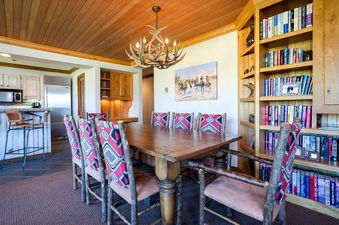 The dining area at Chateau Chamonix CX121 showcases a large wooden table with eight chairs, a unique antler chandelier, and a built-in bookshelf filled with books, all under a wood-paneled ceiling.