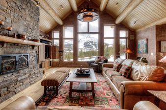 A spacious, rustic-chic living room with a large stone fireplace, leather sofas, a patterned rug, and expansive windows offering stunning views of snow-covered mountains.