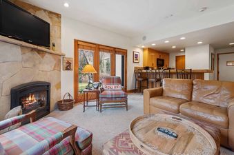 A cozy living room at The Charter E0105 with a stone fireplace, plaid armchair, and a view into the kitchen area with a breakfast bar.