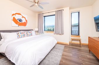 The bedroom at Basecamp Luxe is spacious and bright, featuring a large bed with white linens and a colorful accent pillow, a ceiling fan, and large windows offering views of the surrounding mountains and trees.