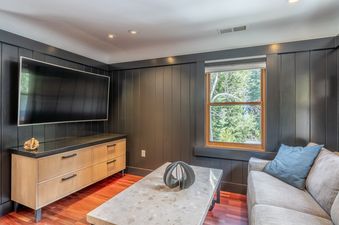 A cozy entertainment room with dark paneled walls, a large TV, a wooden console, a comfortable sofa, and a window offering a view of green trees.