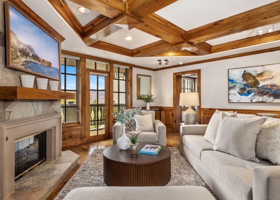 A cozy and elegant living room at Chateau Beaver Creek Residence 309, featuring a stone fireplace, wood-beamed ceiling, comfortable seating, and large windows with an outdoor view.