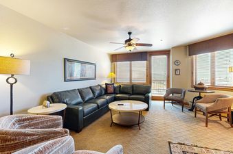 A spacious living room features a dark leather sectional sofa, a floor lamp, a round coffee table, and patterned armchairs, all set on a light brown patterned carpet.