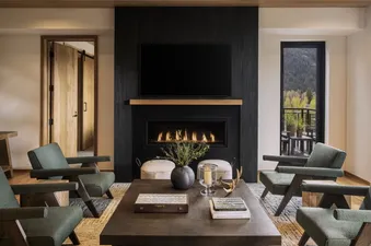 A luxurious living room in The Sylvan Lodge Two Bedroom Suite features a black fireplace, a television, four green armchairs, and a large coffee table, with a stunning mountain view from the window.