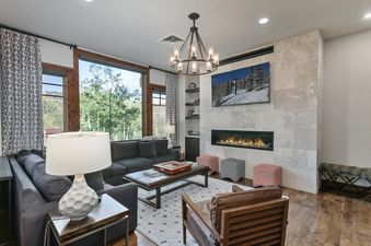 A modern and luxurious living room at Silver Strike Lodge 407 with a gray sectional sofa, a leather armchair, a contemporary coffee table, a geometric rug, a chandelier, and a flat-screen TV above a fireplace, all set against a backdrop of large windows w