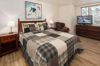 A cozy bedroom at Tamarack Townhomes 34 featuring a queen bed with plaid bedding, wooden furniture, and a wall painting of a skier.