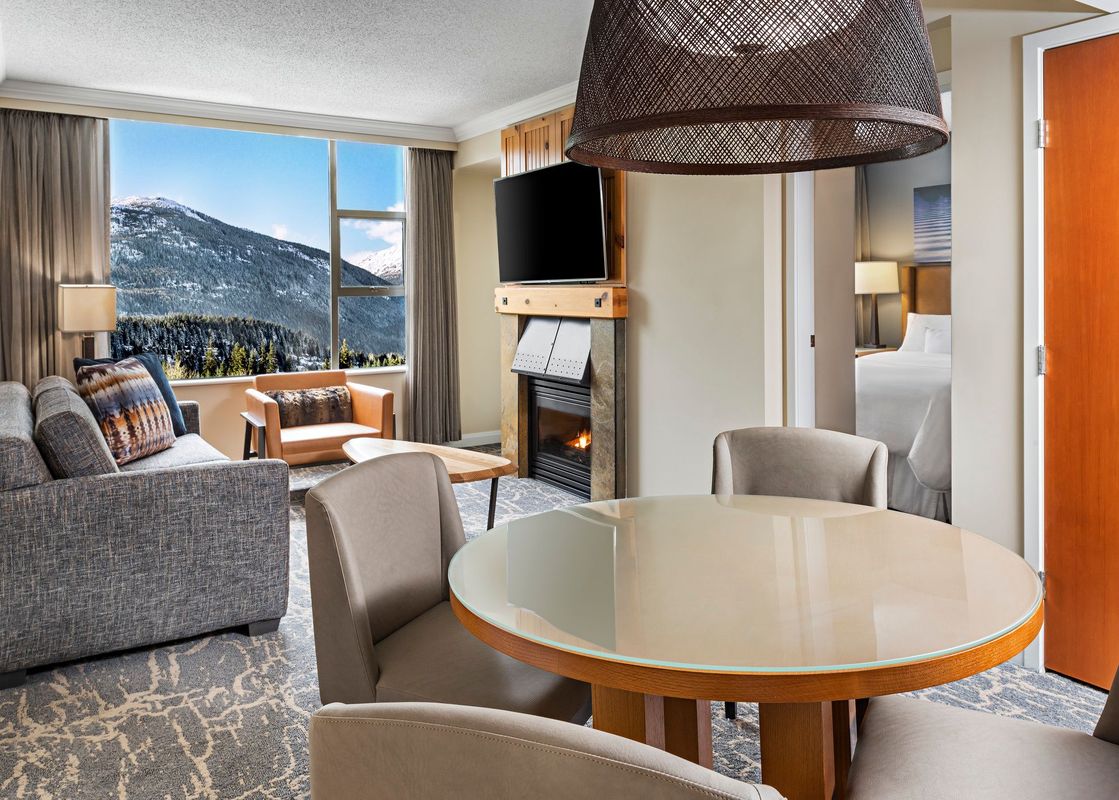 A luxurious living area in The Westin Resort One Bedroom Mountain View features a cozy fireplace, comfortable seating, a dining table, and a large window offering stunning mountain views.