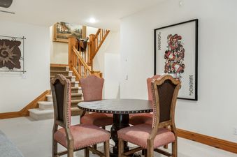 A round wooden game table with four upholstered high-back chairs in the basement of Holden Estate, next to a staircase.