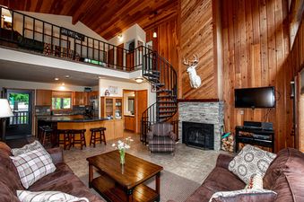 The spacious living room at The Cedar Nest features a stone fireplace, brown leather sofas, and a rustic wooden coffee table, all under a high wooden ceiling with an open-concept kitchen and a spiral staircase leading to a loft.