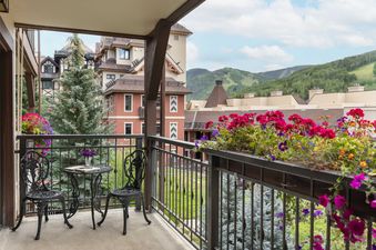 A private balcony with an ornate black wrought-iron table and chairs, colorful flower planters, and a scenic view of alpine buildings and mountains.