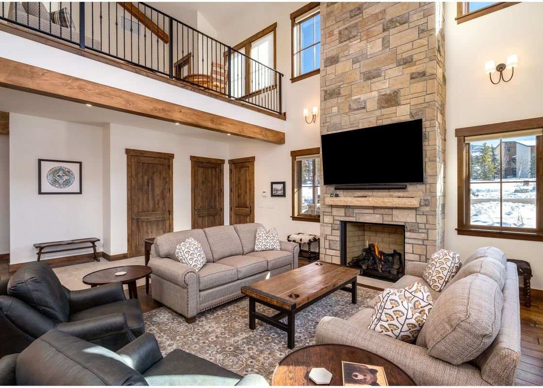 The spacious, sun-drenched living room of The Bear and Moose Lodge featuring a tall stone fireplace, comfortable seating, and a loft overhead.