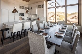 A modern dining room and kitchen with a long dark wooden dining table set for guests and an adjacent kitchen island with white bar stools.