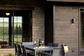 An inviting outdoor dining area at the Northern Hideaway features a dark grey table set with wine and snacks on a flagstone patio, surrounded by comfortable chairs and a stylish, modern house exterior.