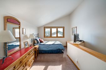 A cozy bedroom at The Pines Condominiums A304, featuring a bed with blue bedding, a large window with snowy views, and a wooden dresser with a red top and mirror.